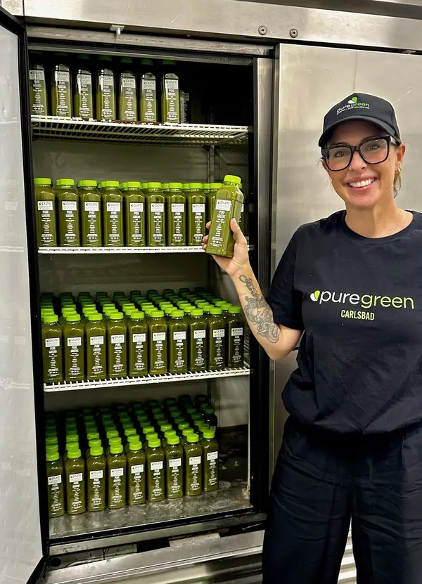 Pure Green Carlsbad team member holding a cold-pressed juice by the stocked juice fridge