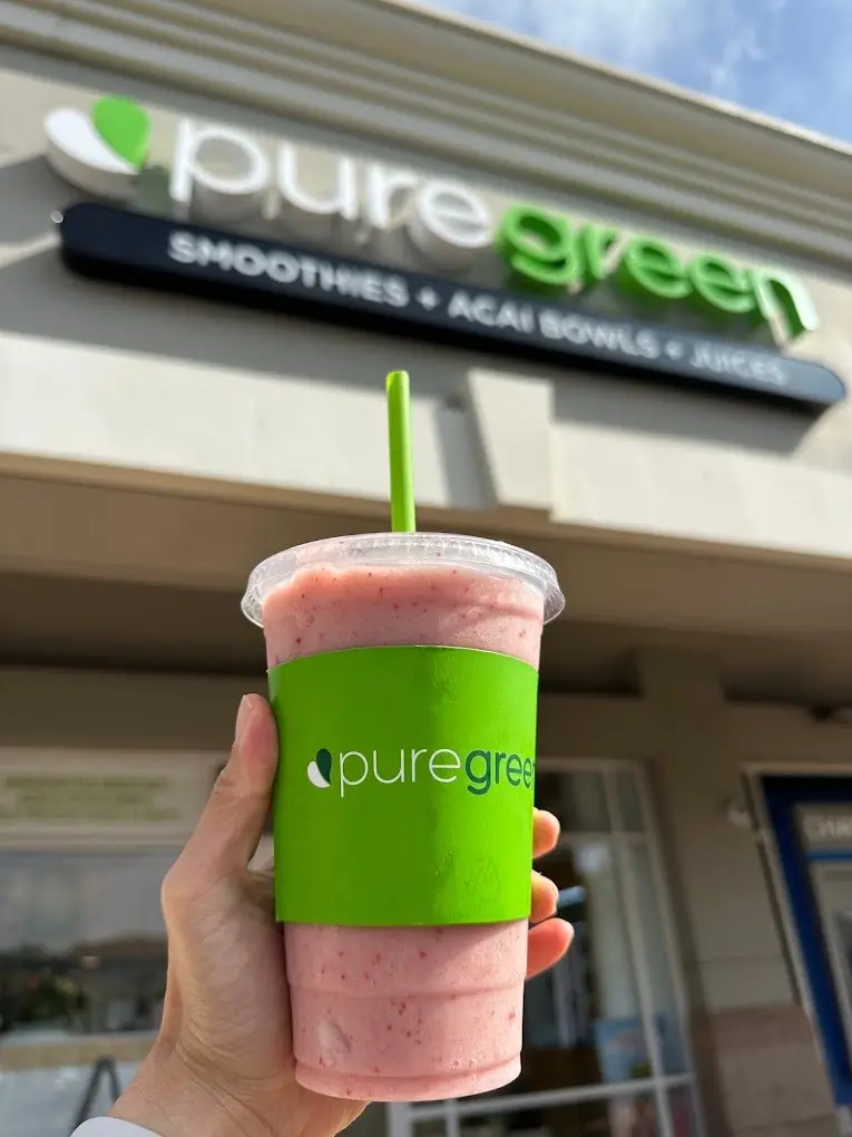 A pink Pure Green smoothie held up in front of the Carmel Valley storefront sign