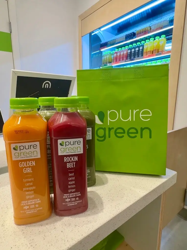 Bottles of Golden Girl and Rockin Beet cold-pressed juice in front of a green Pure Green bag and the juice fridge at the Hoboken store
