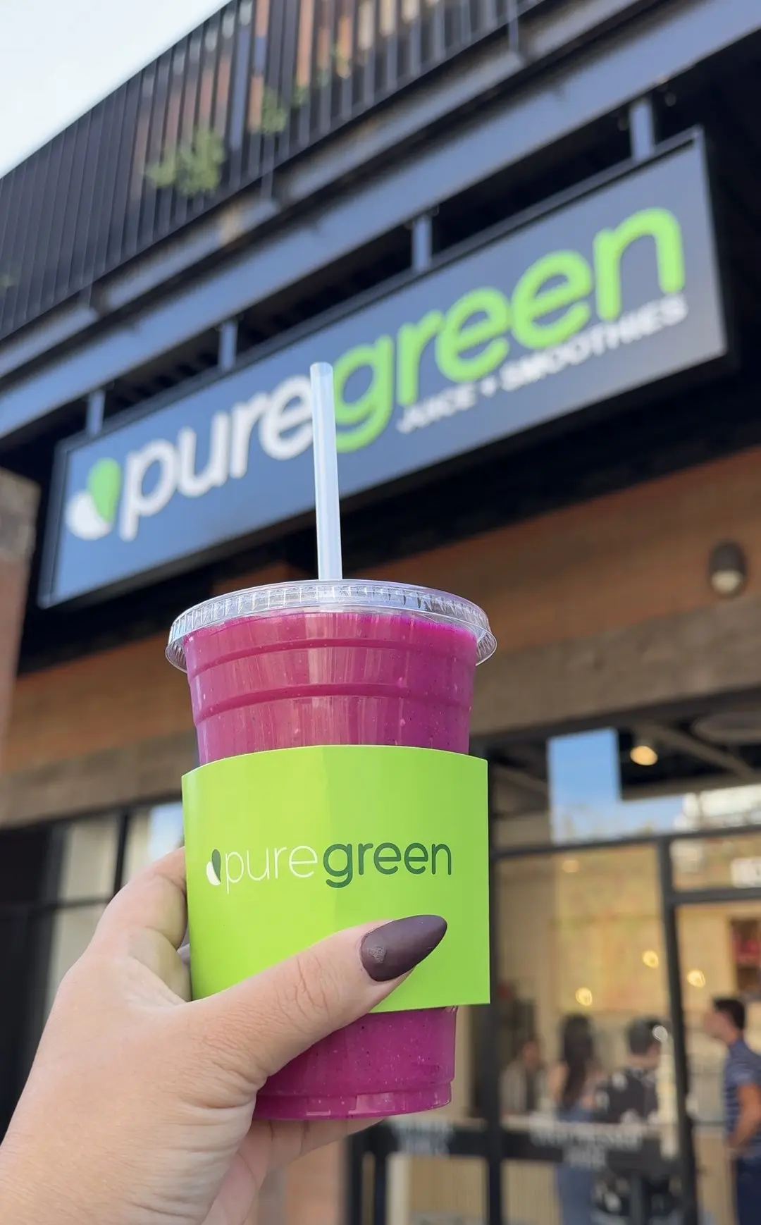 A pink Pure Green smoothie held in front of the Tempe storefront sign on Mill Avenue