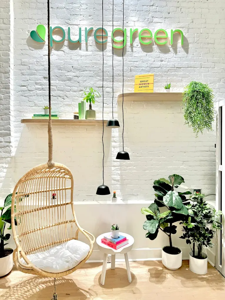 The Pure Green wall logo on white brick above a rattan hanging chair and plants inside the Birmingham store