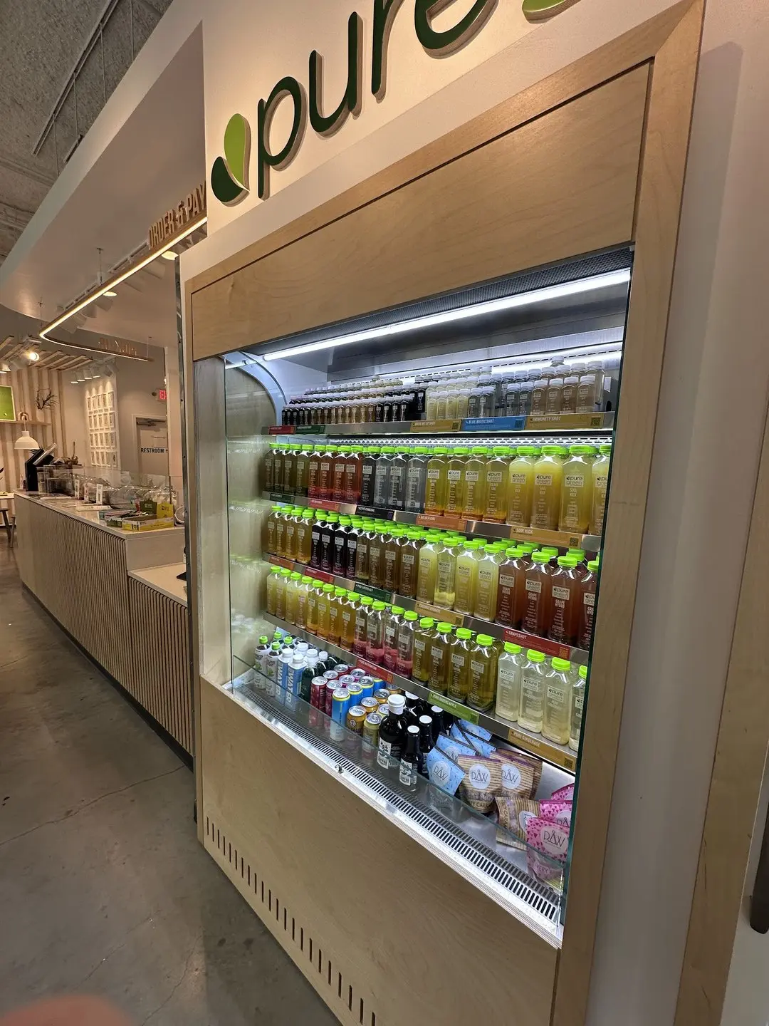 The stocked cold-pressed juice fridge beneath the Pure Green wall logo at Pure Green Capitol Square in Madison