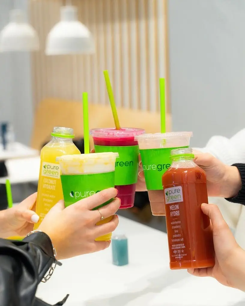 Four hands raising Pure Green cups and cold-pressed juice bottles over a table in the store