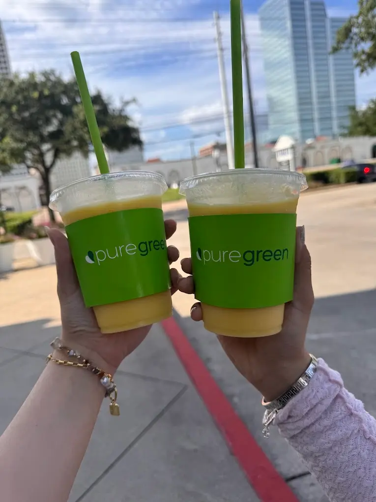 Two smoothies held up outside Pure Green West University with the Houston skyline behind