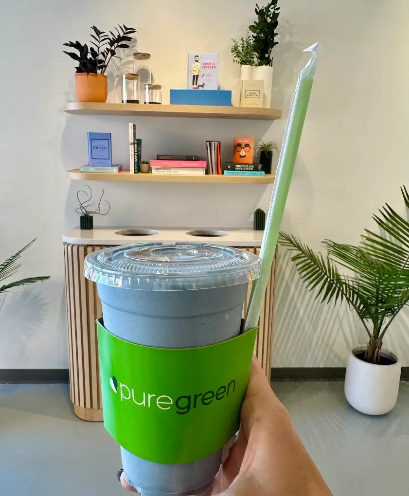 A blue smoothie in a Pure Green sleeve held in front of the book and plant shelves at Pure Green Knoxville