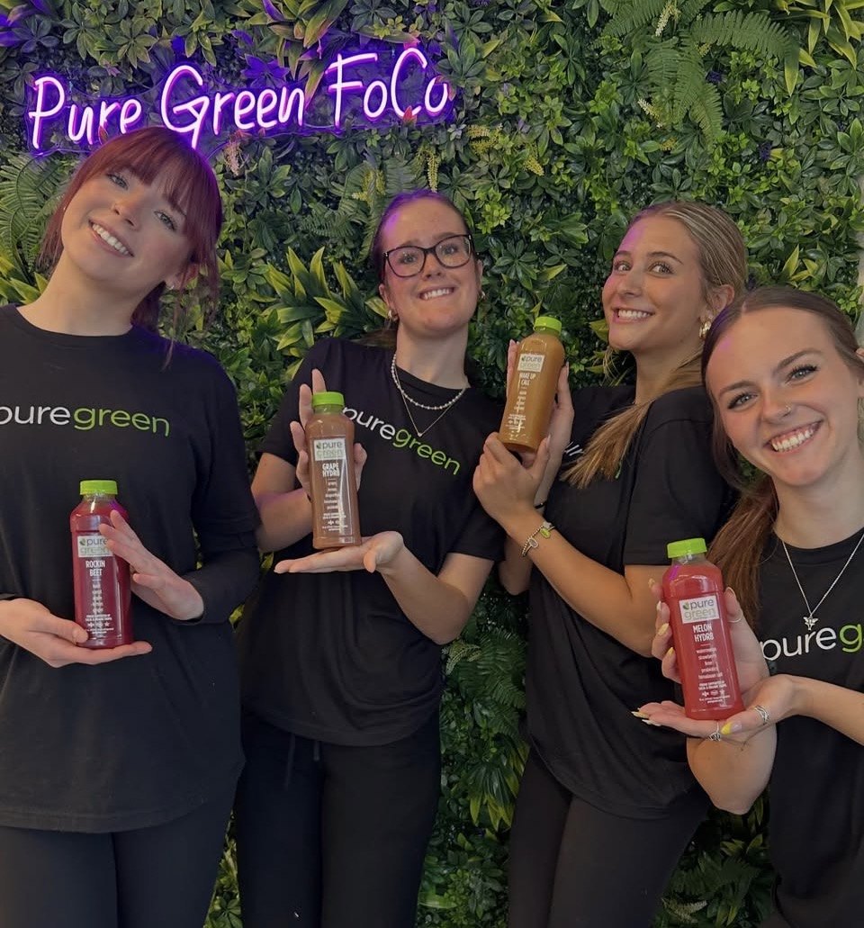 The Pure Green Fort Collins team holding cold-pressed juices by the greenery wall